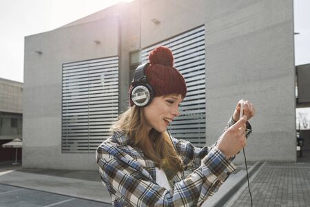 a young woman, wearing a beanie and headphones, that is using a music application in her smart phone device while in an urban, modern city architecture environmentの写真素材