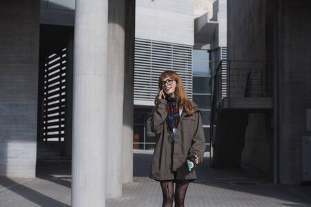 a bohemian style woman with red hair is talking on the phone, in a modern architecture environment while holding a cup of coffeeの写真素材