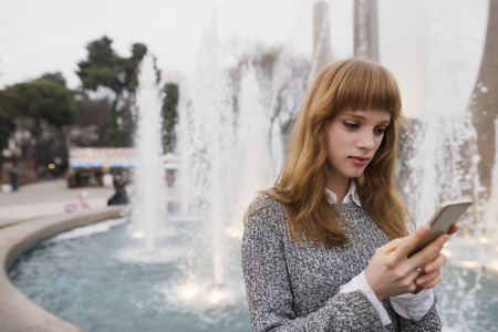 a modern woman with red hair is using an application in her smart phone device to send a text message in front of a fountain in an urban park, at city environmentの写真素材