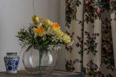 plastic flowers in a vase in a room with a old curtainの写真素材