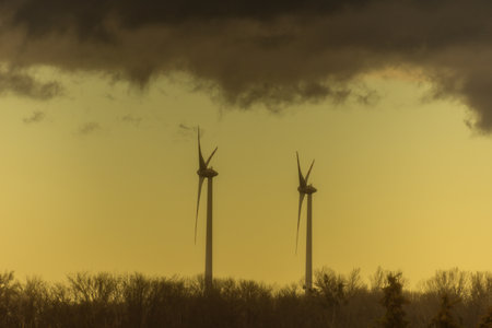 two windmills in a flat landscape with dark rainclouds and yellow color at the skyの写真素材
