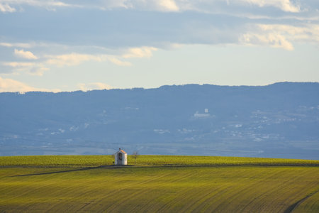 wayside shrine between green fields and a landscape with houses and forest in the backgroundの写真素材