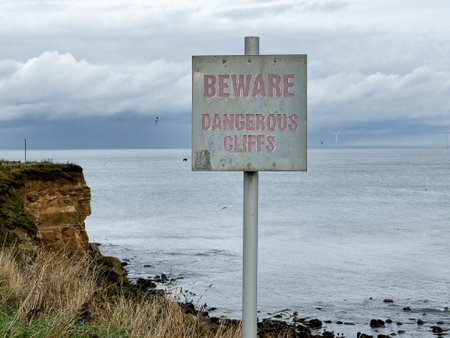 White sign, signpost or post with red letters saying Beware, dangerous cliffs. In front of eroded cliffs and and sea at a beach. In England, UKの写真素材