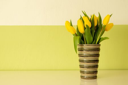yellow tulips in a  flower vase on a white board, white and green wall as background
