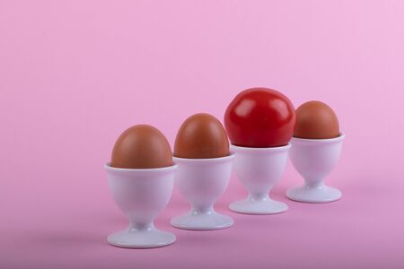 three brown eggs and a tomato in white eggs cups before pink background, close-upの写真素材