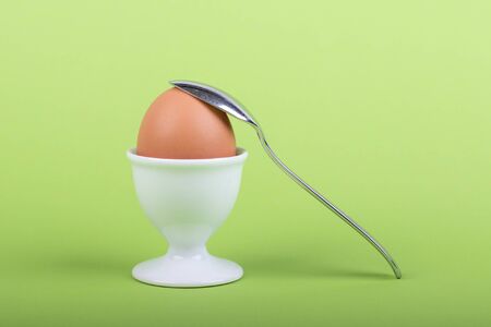 one brown egg in white egg cup before green background, a teaspoon is laying on it, close-upの写真素材