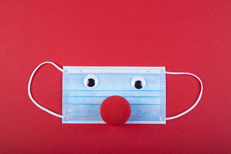 a red foam nose lies under a face mask, symbol for the restriction to carnival due to corona virus, red backgroundの写真素材