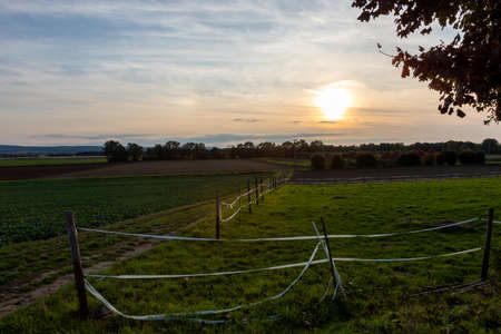 View over farmland at sunset, field with fence in the foreground, dirt road between the fields, cloudy skyの写真素材
