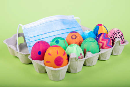 painted eggs with easter motives in an egg carton, face mask behin as symbol for impact of corona...