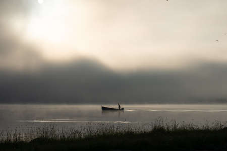 An early morning at Lac de Joux, Switzerland: A lone fisherman on a boat, fog is still hiding the rising sunの写真素材