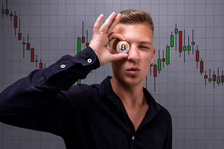 Young guy holds bitcoin in his hand. Cryptocurrency market graph on the backgroundの写真素材