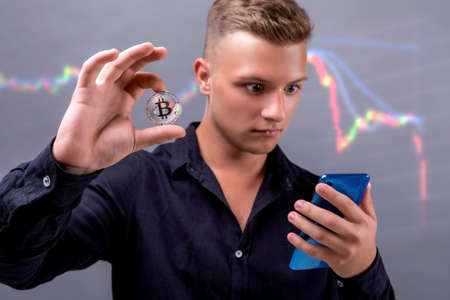 Young guy holds bitcoin in his hand. Cryptocurrency market graph on the backgroundの写真素材