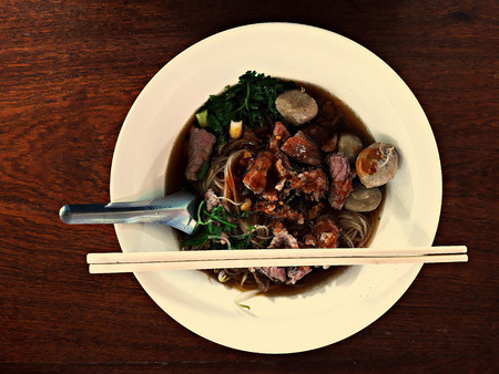 Noodles, meat ball and fresh meat, soup in a white bowl on a wooden table, use a spoon and...