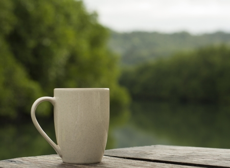 A white cup of coffee is placed on the floor of an old wooden table with a natural backdrop and a blur.の写真素材