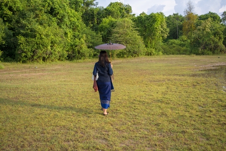Thai women in blue sarongs are walking past the grass and have a backdrop of forest.の写真素材