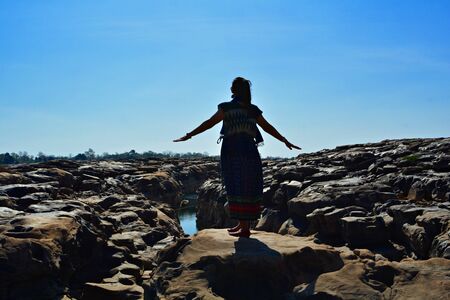 A woman standing with arms stretched on a cliff With a small stream at the bottom (Silhouette photography)の写真素材