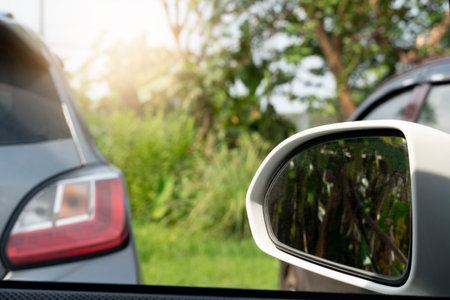 Mirror wing of white car driving on the road. Pass through other car in the parking lot. Parking area with green grass and trees.の写真素材