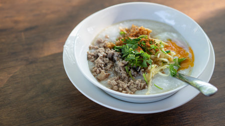 Minced Pork Congee with Egg on white bowl. Spoons are placed and plates are supported on a wooden table top.の写真素材