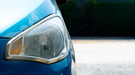 Headlights of bue car. with sunlight casting a radiant glow on the hood. The background depicts a blurred parking lot and dark green trees.の写真素材