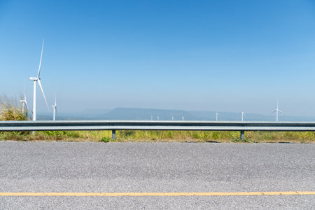 Horizontal view asphalt road with guard lens. Wind turbines tower over a lush forest. Background of mountain far away under blue sky.の写真素材