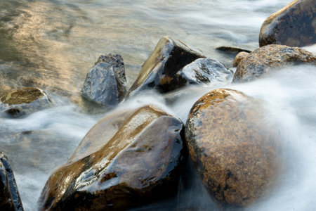 Shows a close-up of a riverbed with smooth  rounded stones. Water is flowing gently over the rocks creating a blurred and ethereal effect.の写真素材