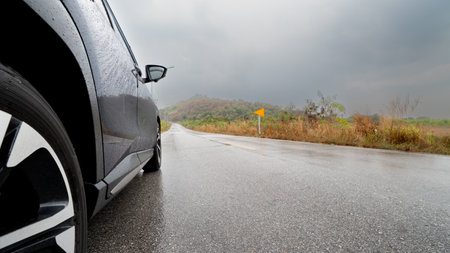Side of car driving on a wet asphalt road under an overcast sky. A blurred landscape of fields and...