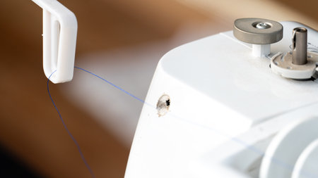 Top of a white sewing or embroidery machine. A spool of blue thread is feeding into the machine from...