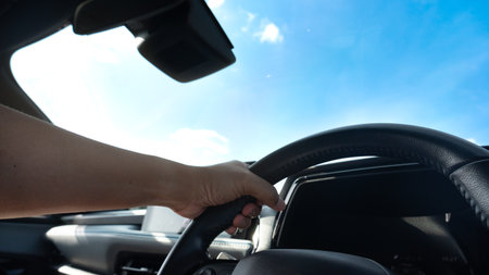 Hand on steering wheel of car on blue sky and white clouds background. Single hand control steering...