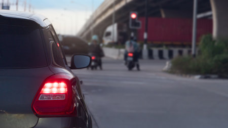 Rear side of dark car turn on brake light on the road. Car park at the intersection with a red traffic light. There are cars going straight across. Under the overpass at evening.の写真素材