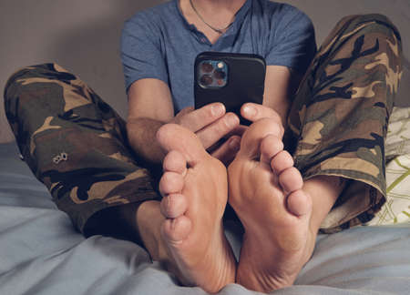 Man barefoot on bed holding mobile phone in hands
