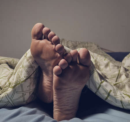 Man lying barefoot in bed under blanketの写真素材