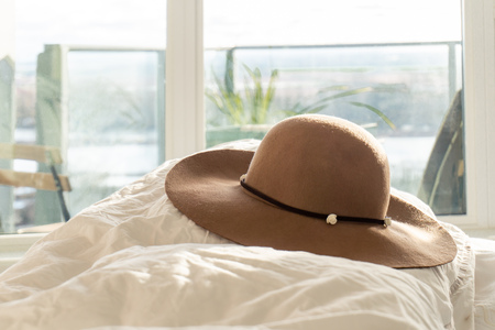 Beige woman's round sun hat with floral trim and wide brim, on a fluffy comforter in a bedroom, with balcony in the background. Depicting holidays, relaxation, lifestyle and fashion style.の写真素材