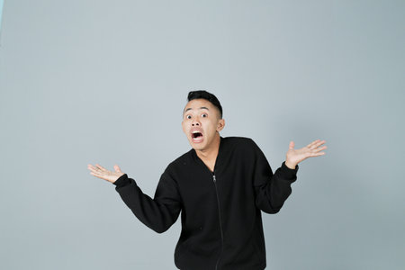 a young man wearing a black coat is showing a gesture of ignorance, isolated on a gray backgroundの写真素材