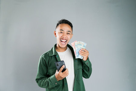 a young asian man is holding a smartphone and some moneyの写真素材