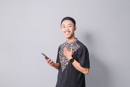 young asian muslim man is holding smartphone while smiling, isolated on gray backgroundの写真素材