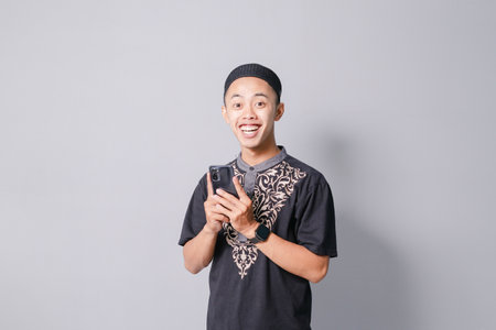 young asian muslim man is holding smartphone while smiling, isolated on gray backgroundの写真素材