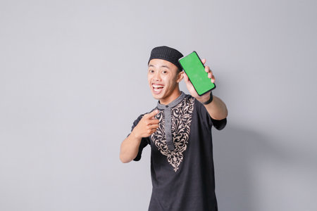 young asian muslim man is holding smartphone while smiling, isolated on gray backgroundの写真素材