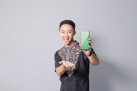 young asian muslim man is holding smartphone while smiling, isolated on gray backgroundの写真素材