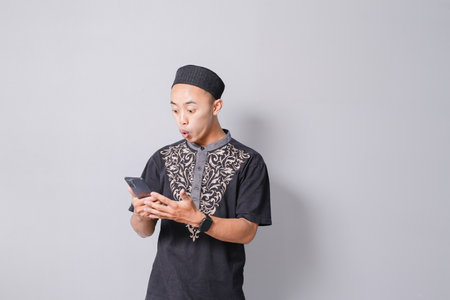 young asian muslim man is holding smartphone while smiling, isolated on gray backgroundの写真素材