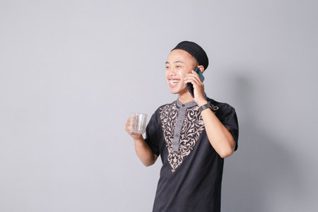 Young Asian Muslim man holding a smartphone while carrying a glass of drinking water, isolated on a gray backgroundの写真素材