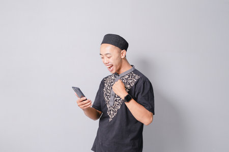 young asian muslim man is holding smartphone while smiling, isolated on gray backgroundの写真素材