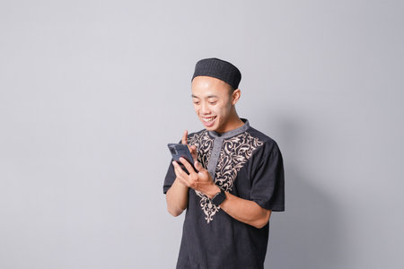 young asian muslim man is holding smartphone while smiling, isolated on gray backgroundの写真素材