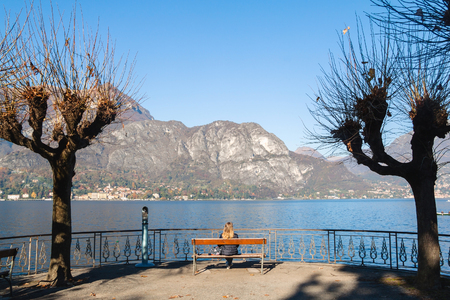 Young girl sitting on a bench looking at lake and mountains. Como Lake, Italy.の写真素材