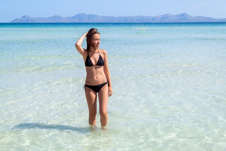 Young woman in bikini standing with her hand over head enjoying hot summer day on the beautiful beach of Alcudia, Mallorca, Spain.の写真素材