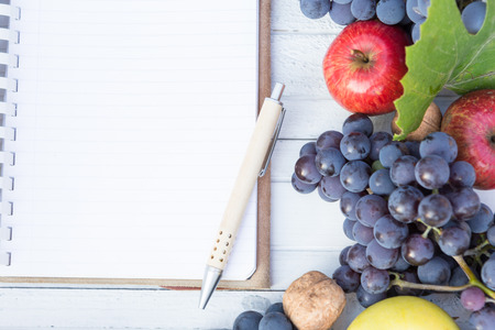 Blank notebook with wooden pen and autumn grapes, wallnuts and apples on a white wooden tableの写真素材