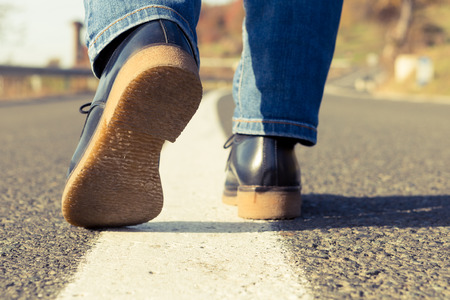 Woman feet walking on the middle of a country side road wearing black leather shoes and jeans on a sunny day of autumn or fallの写真素材