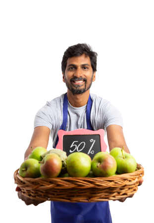 Indian smiling supermarket employee holding organic apple basket with fifty percent discount sign as fresh and healthy concept isolated on whiteの写真素材