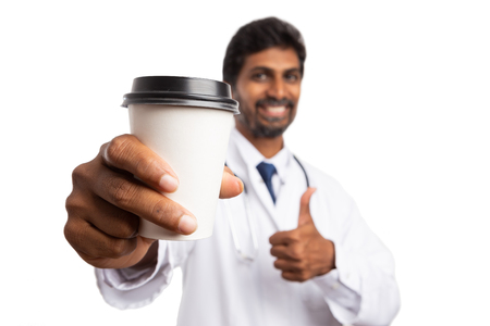 Close-up cup of coffee to-go held by friendly indian medic with thumb up as approve gesture isolated on white backgroundの写真素材
