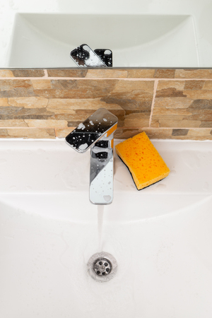 Running water in clear porcelain sink to rinse white foam inside with sponge on the edgeの写真素材