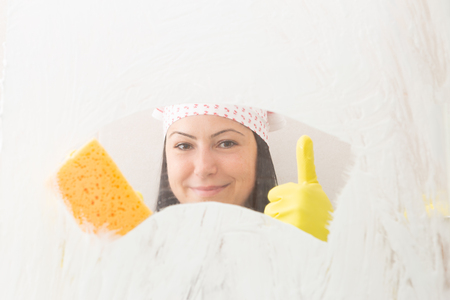 Woman wearing yellow gloves and bandana holding orange sponge in one hand with thumb up smiling from behind shower glassの写真素材